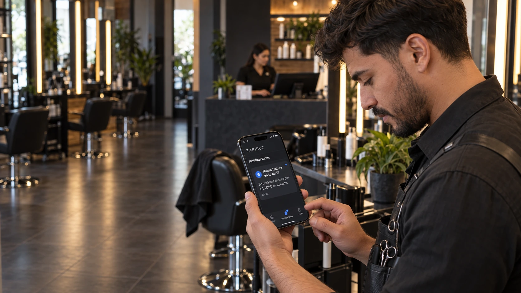 Estilista revisando en su celular el aviso de una nueva factura en TAPIRUZ, con la cajera al fondo.